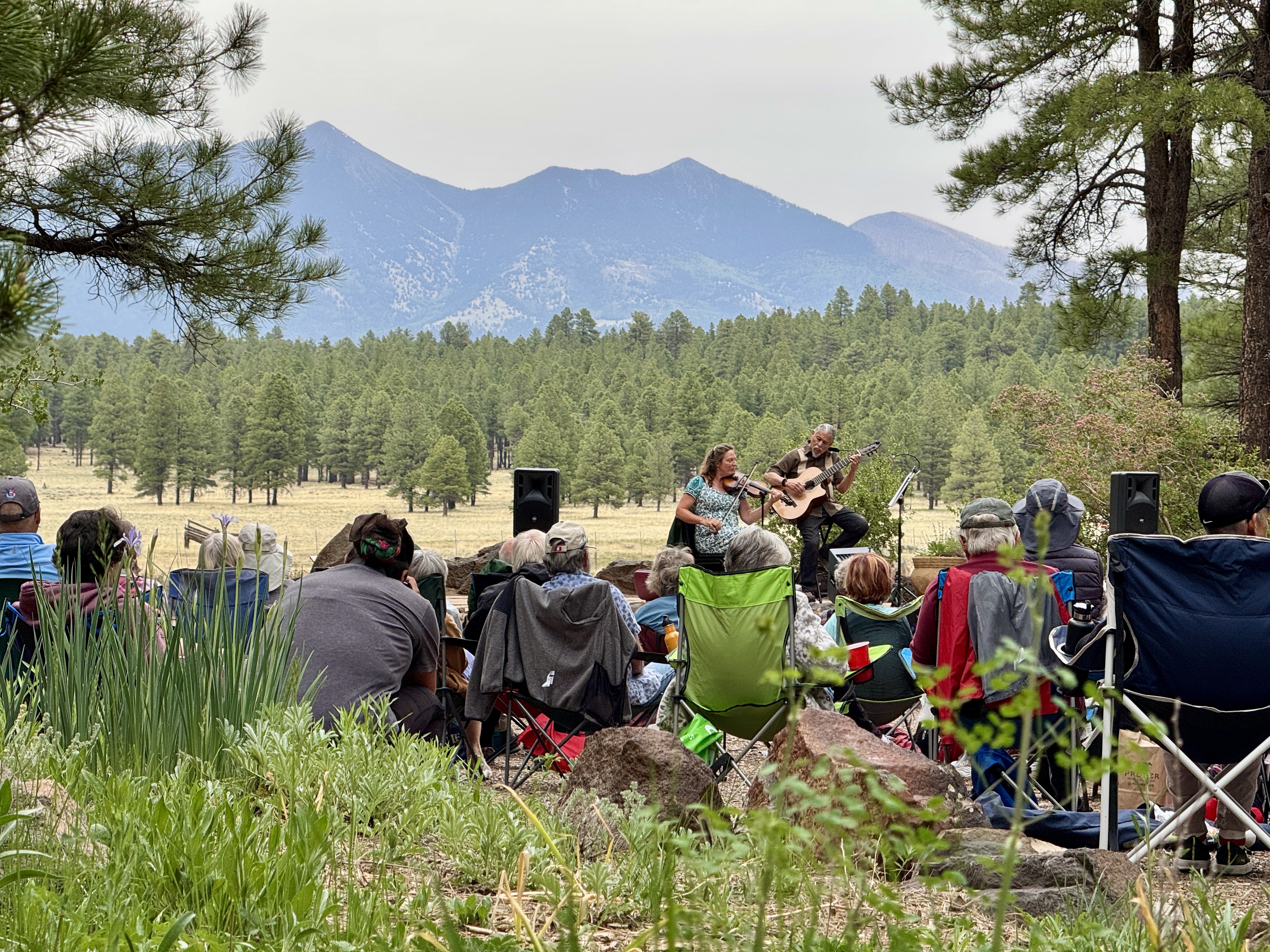 Concert in the field with a mountaintop in the distance, audience in front, presenting a duo, man playing guitar/violin an...